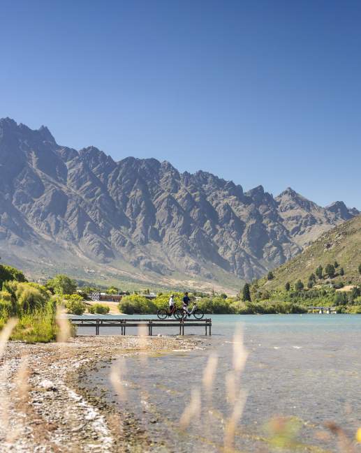 Two people having a rest on a pier looking out over the lake and Remarkables mountains while biking the Frankton Trail