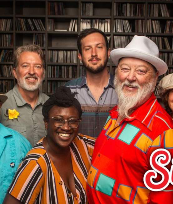 9-piece band poses in front of a bookshelf full of records