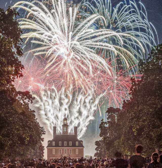 Fireworks illuminate the night sky above a historic building, surrounded by a crowd of spectators. Lush trees frame the scene, enhancing the festive atmosphere.