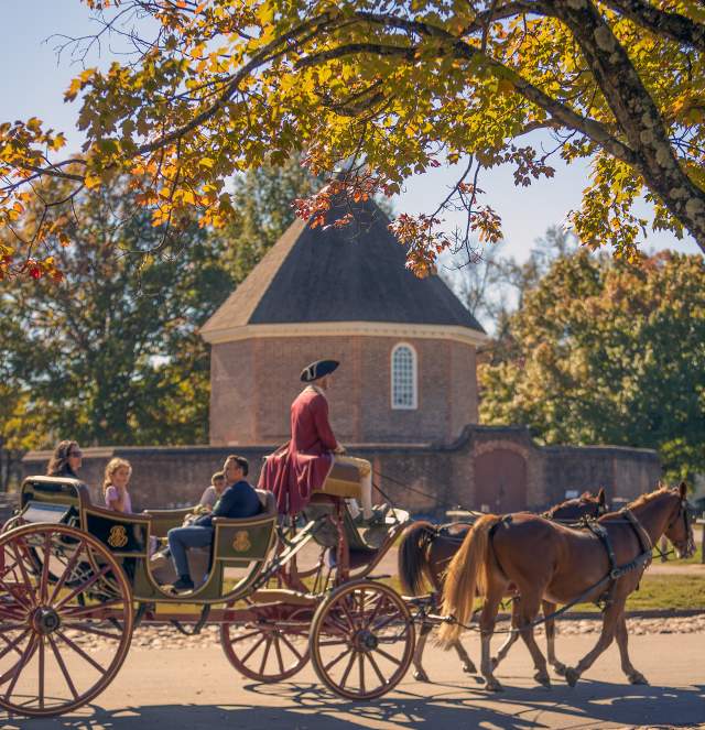 A horse-drawn carriage passes under colorful autumn leaves, with a historic round building in the background, evoking a charming, nostalgic atmosphere.