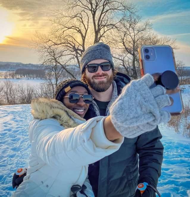 A couple taking a selfie in the winter, surrounded by snow, with the Mississippi River and the sunset behind them.