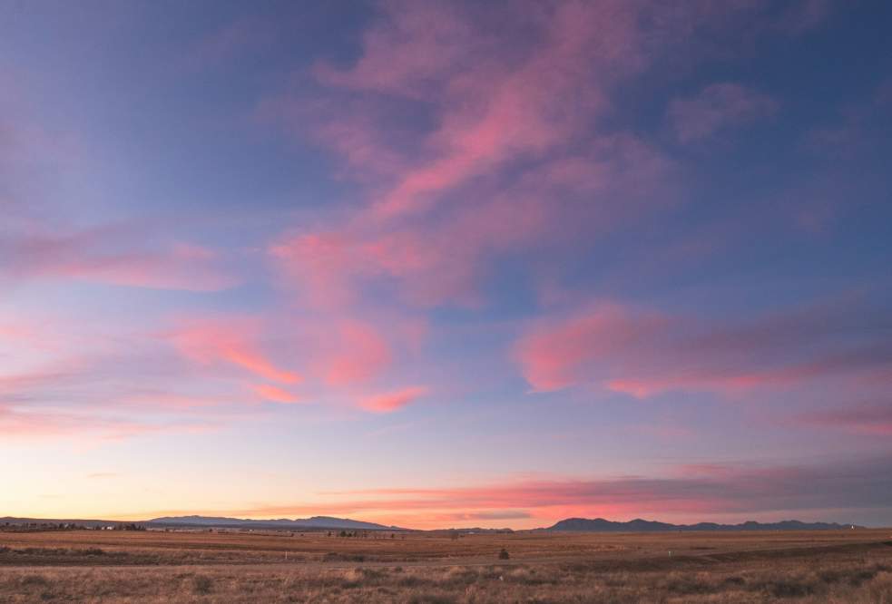 Expansive landscape with a serene sunset sky, featuring pink and purple clouds over a vast, dry plain and distant mountains on the horizon.