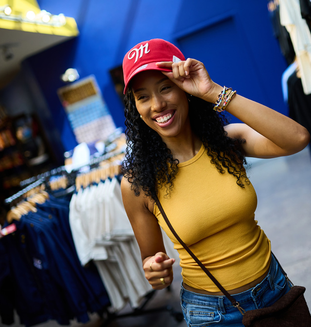 Smiling shopper trying on a red Miller-branded baseball cap inside the Miller Brewery gift shop, surrounded by racks of apparel and branded merchandise.