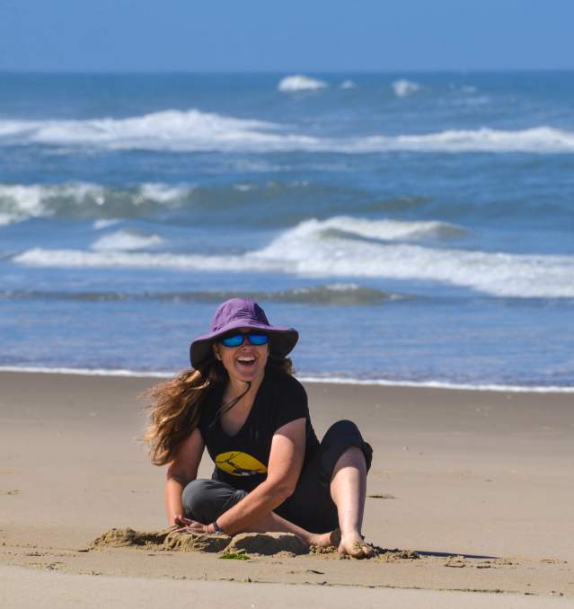 Woman sits building a sand castle on the beach with the ocean behind her. She is smiling under a large, purple brimmed hat.