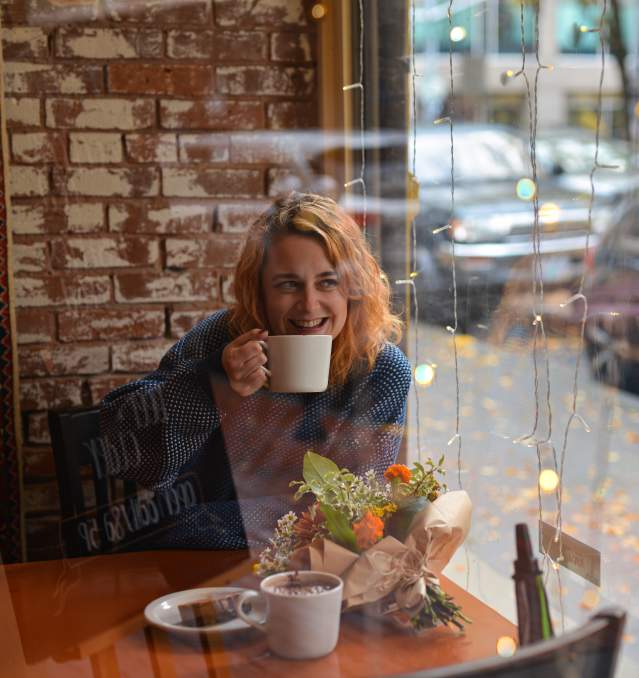 A person smiles with a cup of coffee near a coffee house window