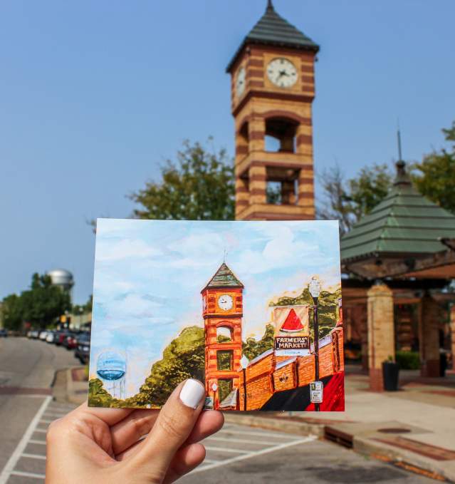 A handpainted postcard showing the downtown Overland Park clocktower is held up in front of the physical clocktower.