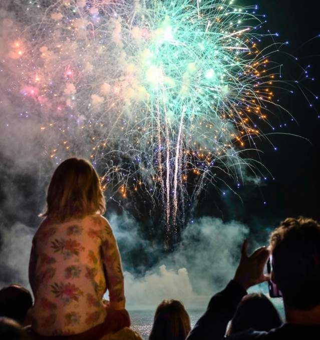 A child sits on their parent's shoulders watching a fireworks show