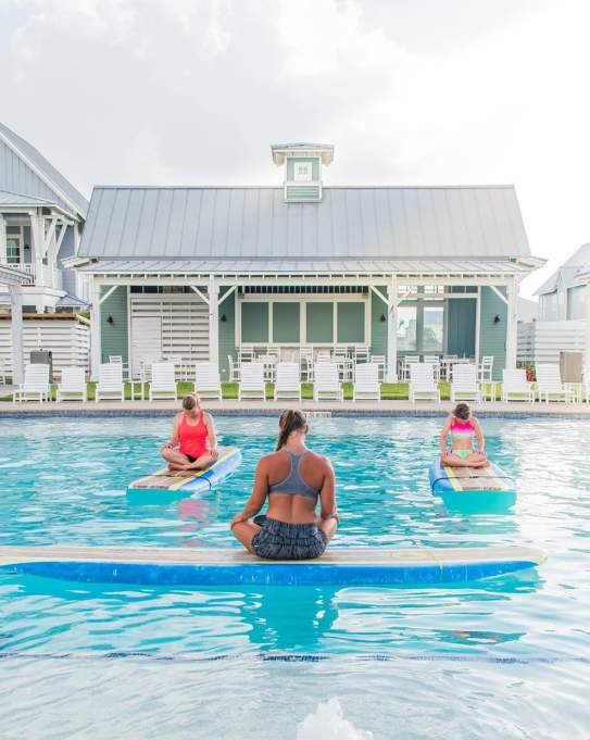 A woman with crossed legs sits atop a surf board floating in a pool, back to the camera. Four women, also with crossed legs, sit atop surf boards facing the instructor.