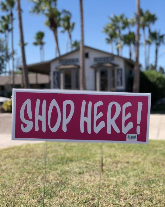 A rectangular pink sign reading "Shop Here!" takes up the middle of the photo. A building surrounded by palm trees is visible in the background.