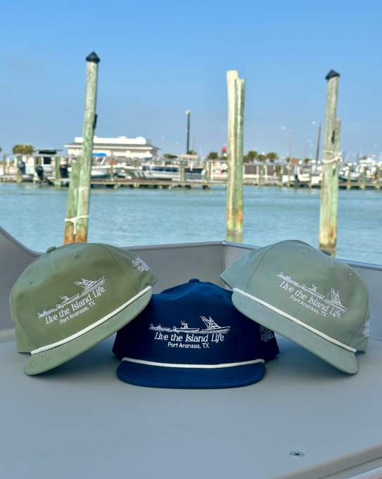Three "Fishing Capital of Texas" hats in green and blue sitting on a table near a sunny harbor.