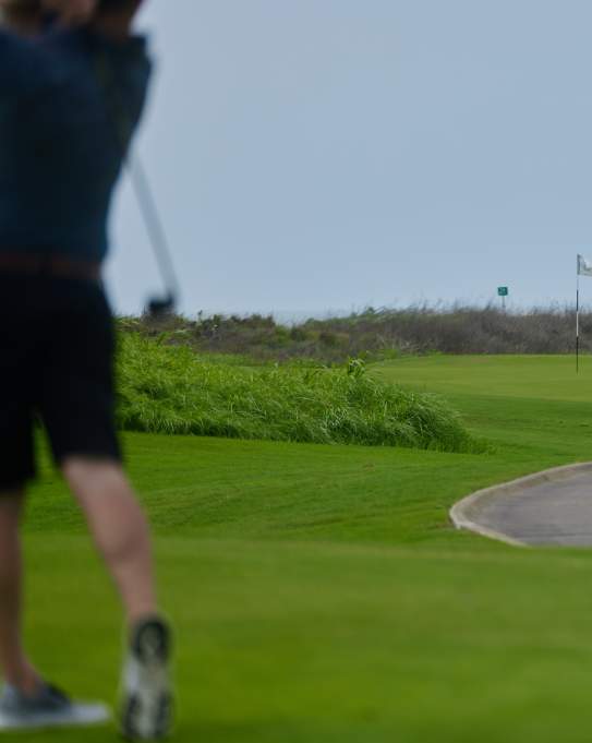 The left side of the photo shows an unfocused man dressed in black golf clothes swinging a club. The rest of the photo is a manicured golf course with a flag in the center and the sea in the background