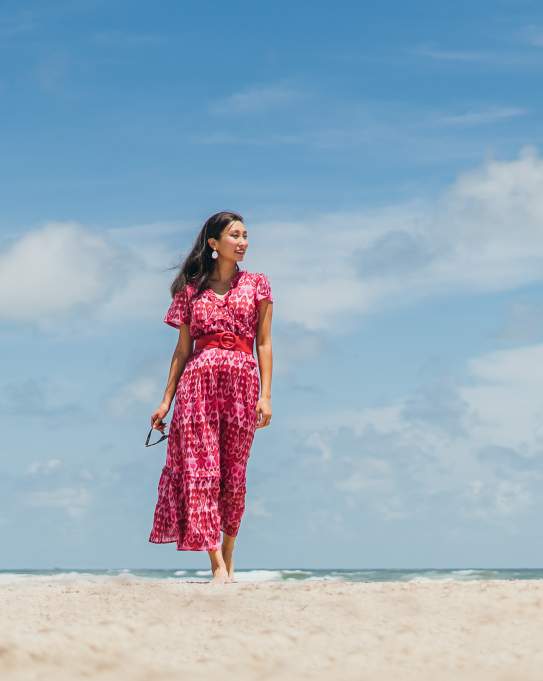 A woman in a pink and red dress walks along the beach