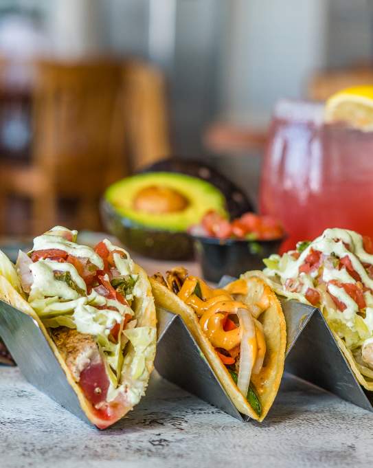A trio of tacos sits on a table surrounded by an avocado, a pink margarita, and other ingredients