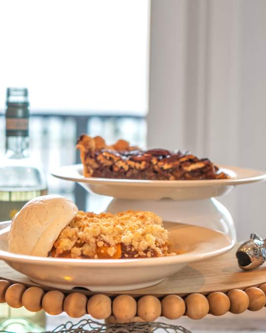 A plate of cobbler with ice cream and a plate of pecan pie sit stacked on an elevated dessert tray. To the left are open bottles and glasses of wine.
