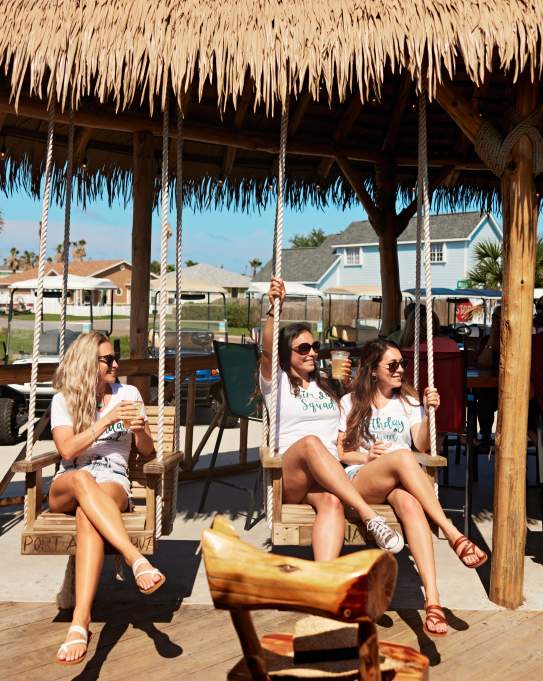 A group of five women wearing matching birthday shirts sit in tiki-style swings