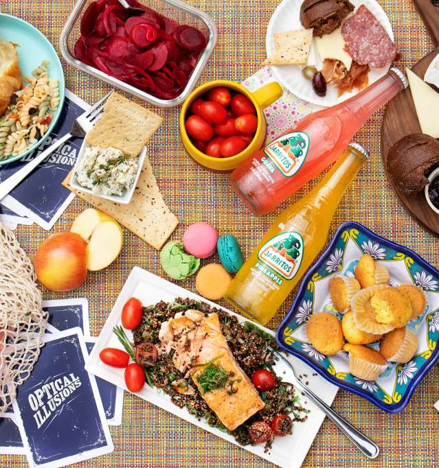 bottled sodas, charcuterie board with meats and cheeses, and various other foods on a table with cards.