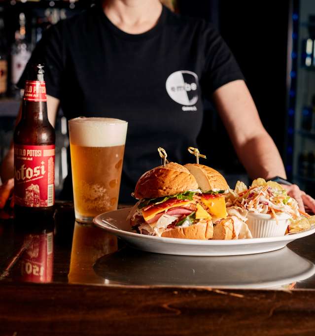 Plate of burgers with a cold, frothy beer on a bar top, with someone standing in the background.