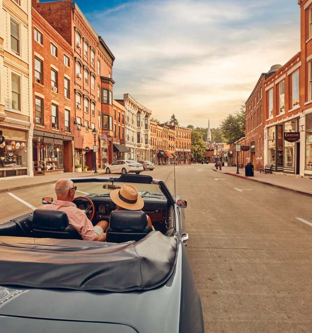 Two people riding in a blue convertible car, down a Main Street of brick buildings.
