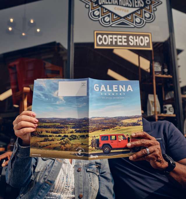 Two people holding a visitors guide, holding it in front of their faces, hands showing. They are sitting in front of a coffee shop.