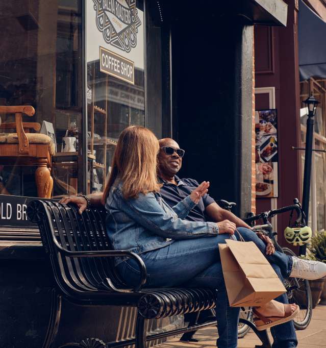 Two people sitting on a bench with a shopping bag, in front of a coffee house.