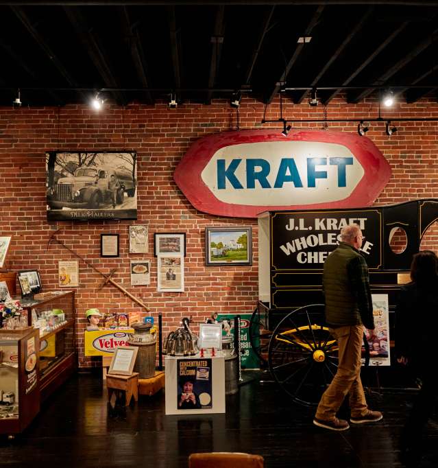 Two people are touring a museum with signs from the past, like Kraft, with a brick wall behind them.