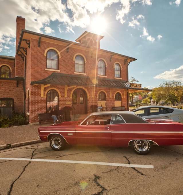 A red, classic car, parked in front of an old, red brick building with the sun shining in the background.