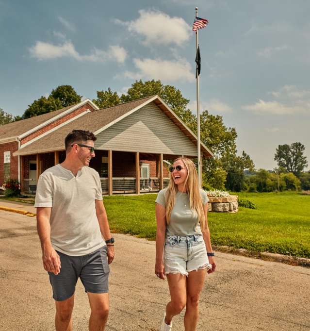 Two people, in summer, walking down a street with a VFW building behind them, and rolling hills.