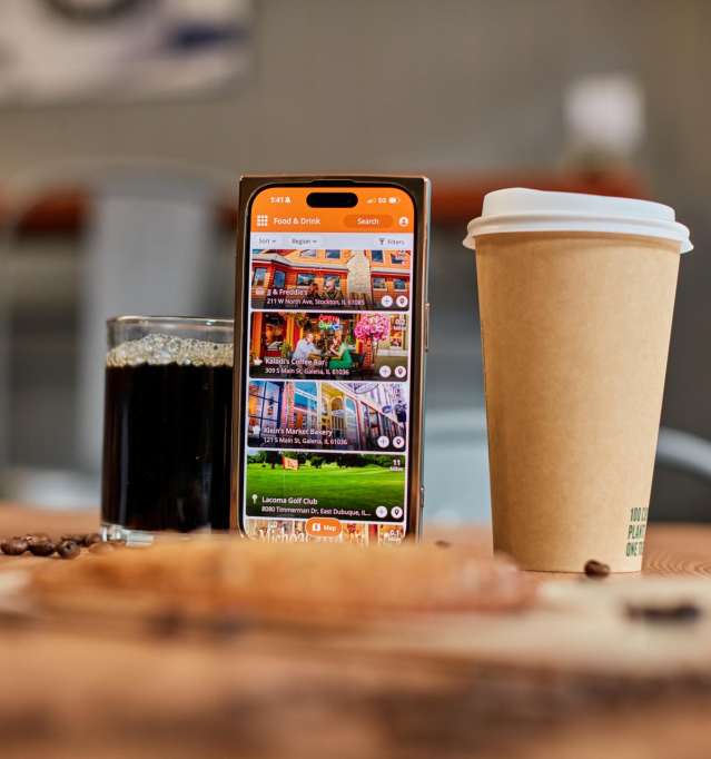 Two cups of coffee, one in a to-go mug, and a cellphone with the Galena Country App pulled up. All set on a table covered in coffee beans and a cookie.