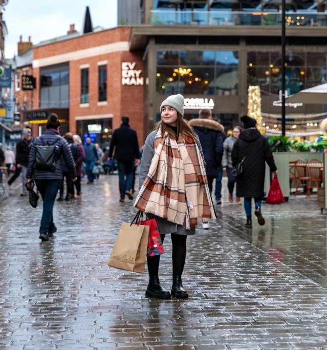 A woman in a scarf is stood in the centre of the Cornhill Quarter in Lincoln, holding shopping bags.