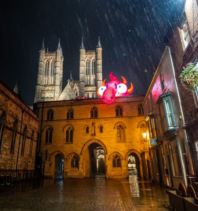 A rainy nighttime shot of Lincoln Cathedral with an inflatable monster on top of a building in the foreground