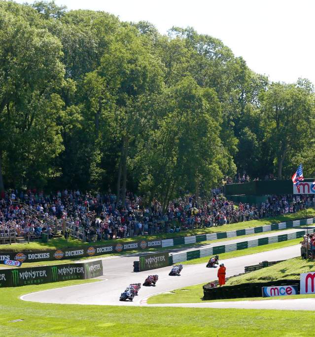 A race track with motorbikes and audience stood around on grass banks