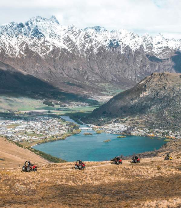 A scenic view of Queenstown, New Zealand, captured from a high vantage point. The image shows snow-capped mountains in the background, a town nestled by a serene lake in the valley, and a group of quad bikes traversing a grassy hill in the foreground.