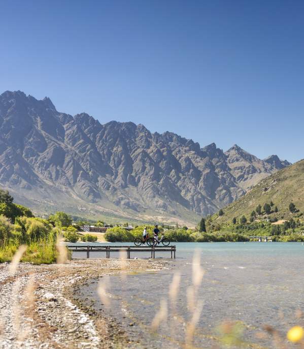 Two people having a rest on a pier looking out over the lake and Remarkables mountains while biking the Frankton Trail