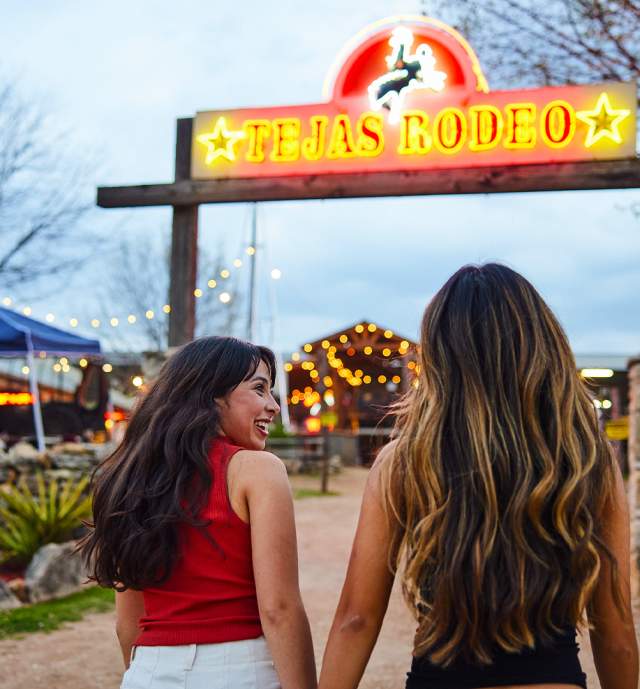 Two girls walking under neon sign that reads "Tejas Rodeo."