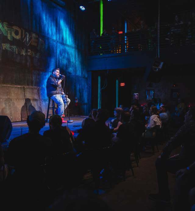 A man sitting on a stool onstage with a mic during a comedy set at Vulcan Gas Company.