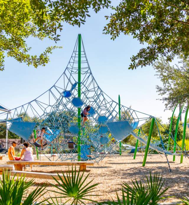 Two children climbing on a netted playscape while their parents watch.