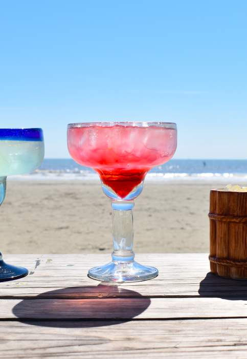 A row of margaritas sitting on a wooden table on the beach.