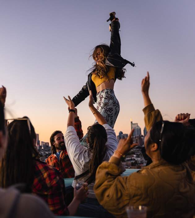 A woman dances with a crowd on a rooftop, overlooking a city skyline.