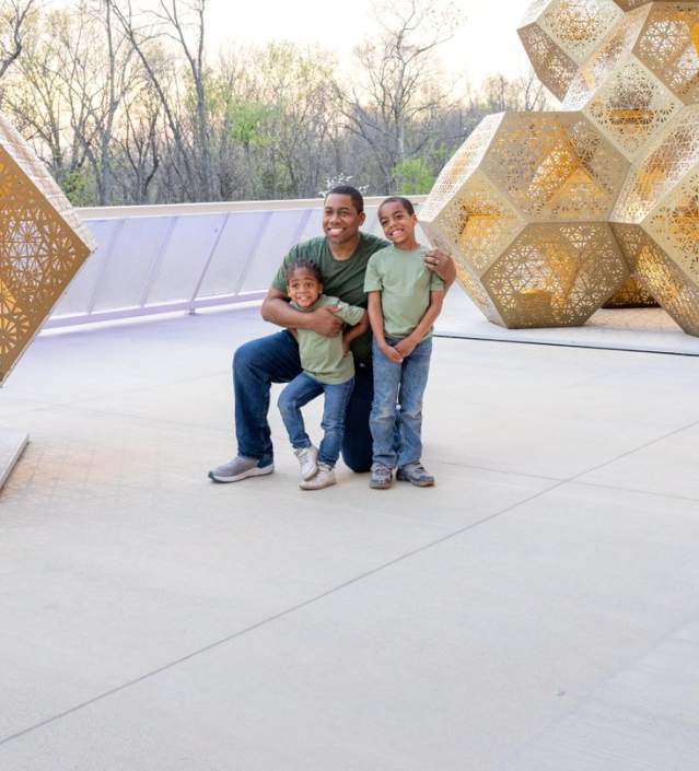 A smiling father kneels and embraces his two young children in front of large, geometric, golden sculptures, part of the HYBYCOZO installation.