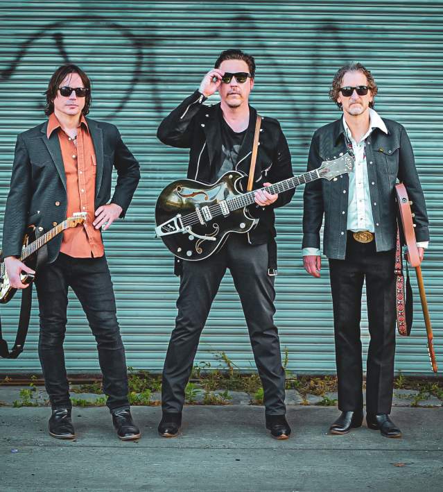 Three-piece band with guitars pose in front of a closed green tagged garage door