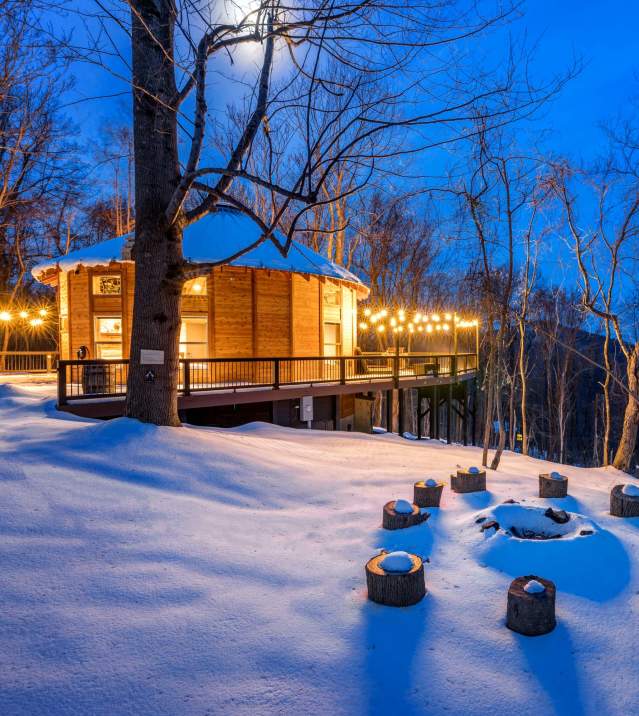 Cozy wooden Shenandoah yurt in snowy Stanley, VA forest at twilight with string lights and fire pit.