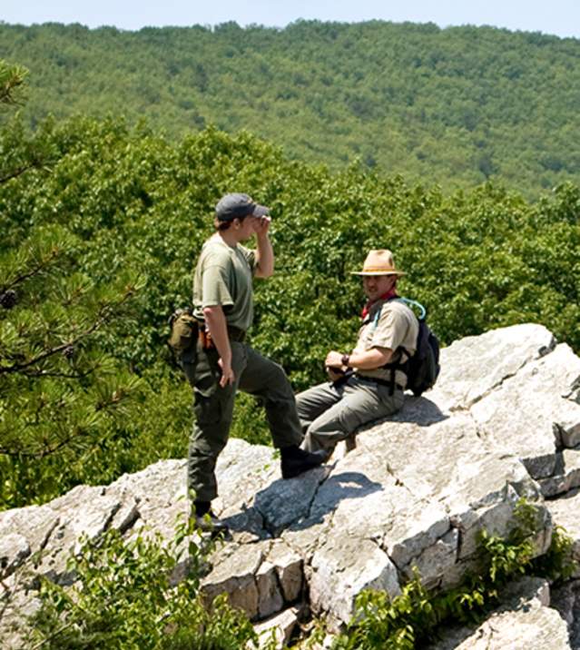 Two men sitting on a rocky outcrop with green hills and a clear sky at Pine Grove Furnace State Park.