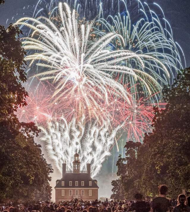 Fireworks illuminate the night sky above a historic building, surrounded by a crowd of spectators. Lush trees frame the scene, enhancing the festive atmosphere.