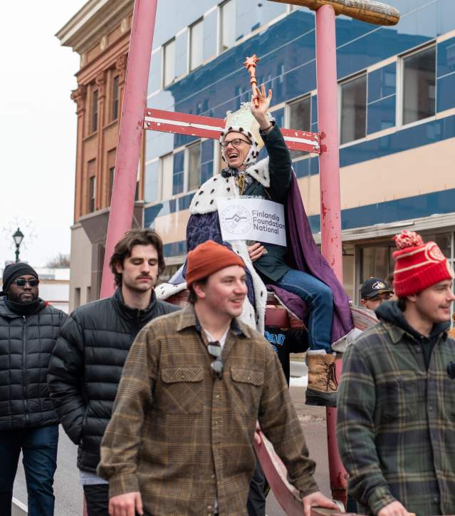a crowned person waves a scepter from atop a kicksled float