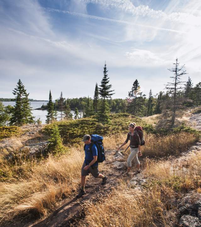 two figures with large backpacks hike across a rocky landscape strewn with grasses and pines. lake superior in the distance
