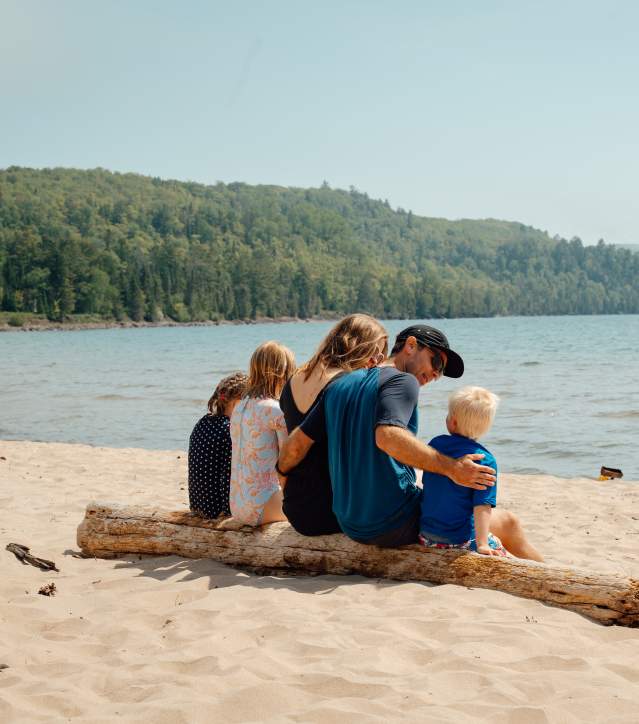 A family sits on a log on a sugar sand beach looking at Lake Superior.