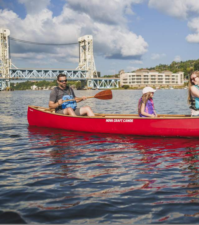 two adults and a child paddle a canoe on gently lapping waters, a lift bridge and summer skies in the background