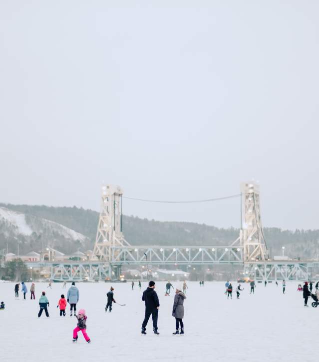 Families skate on the frozen Portage Canal with the Portage Lake Lift Bridge in the background.