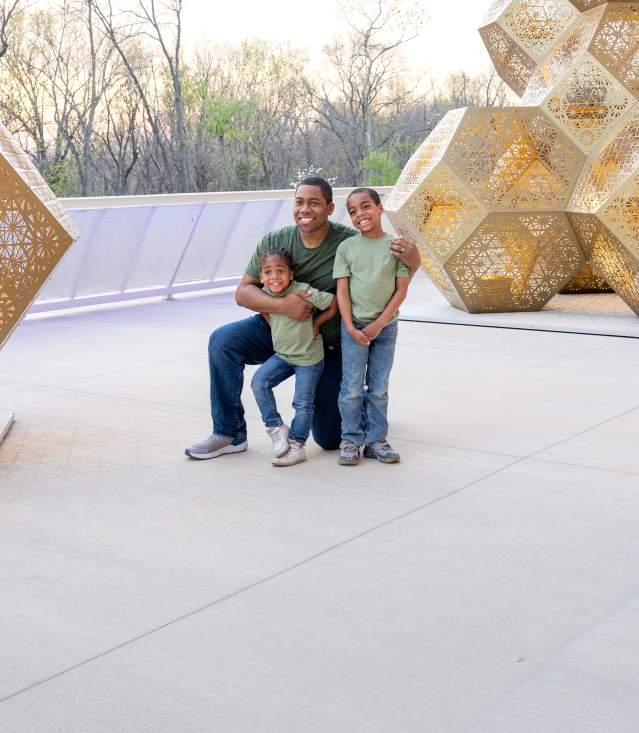 A man and two children pose together in front of large, intricate golden sculptures on a modern outdoor terrace. Trees are visible in the background.