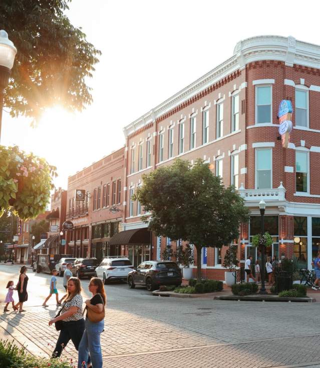 Historic brick buildings line a bustling street, featuring the Walmart Museum. People stroll past shops and outdoor dining areas.